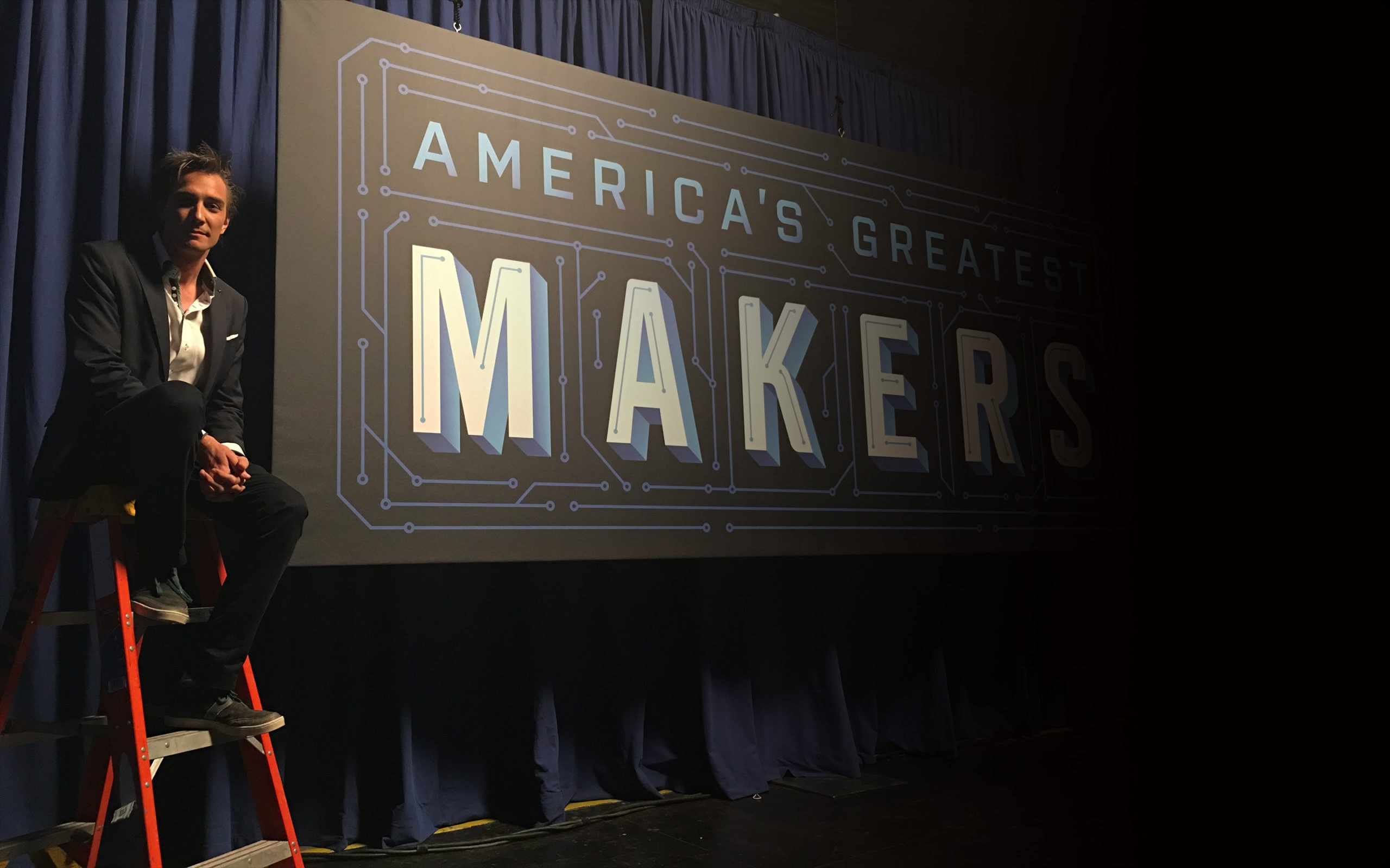 Gray Bright sitting on a ladder in front of the America's Greatest Makers sign