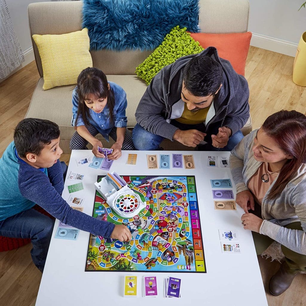Game of Life 2021 'Your Life Your Way' Total Product Offering with game board, money, cards, and spinner, in a lifestyle photoshoot with family. Made by Hasbro Gaming