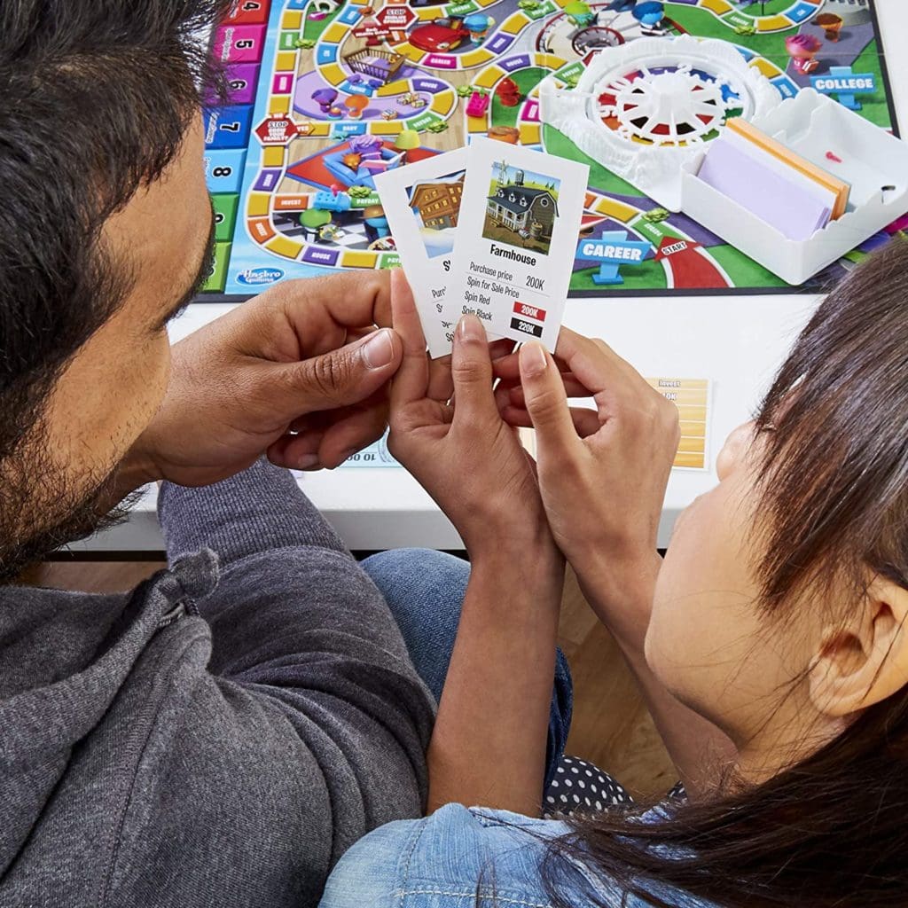Game of Life 2021 'Your Life Your Way' Total Product Offering with game board, money, cards, and spinner, in a lifestyle photoshoot with 2 people. Made by Hasbro Gaming
