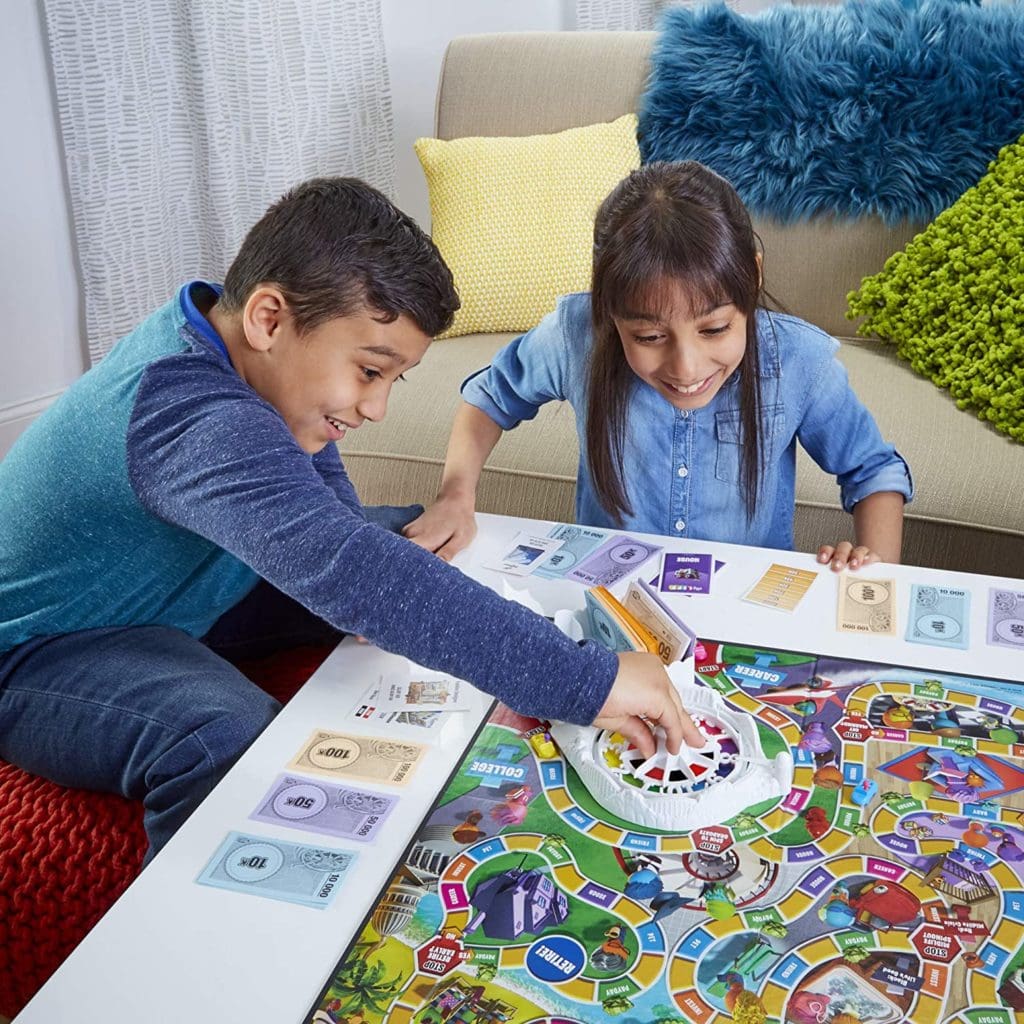 Game of Life 2021 'Your Life Your Way' Total Product Offering with game board, money, cards, and spinner, in a lifestyle photoshoot with 2 kids. Made by Hasbro Gaming