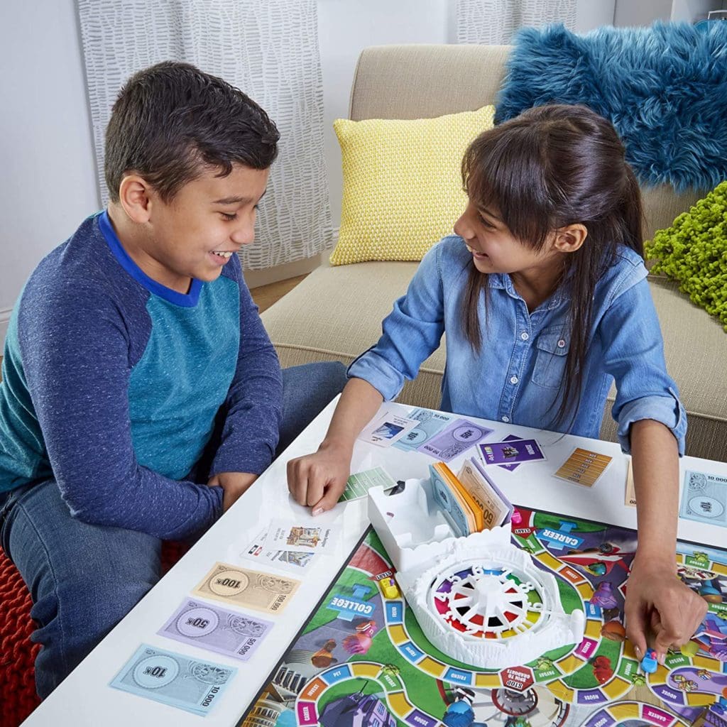 Game of Life 2021 'Your Life Your Way' Total Product Offering with game board, money, cards, and spinner, in a lifestyle photoshoot with 2 kids. Made by Hasbro Gaming