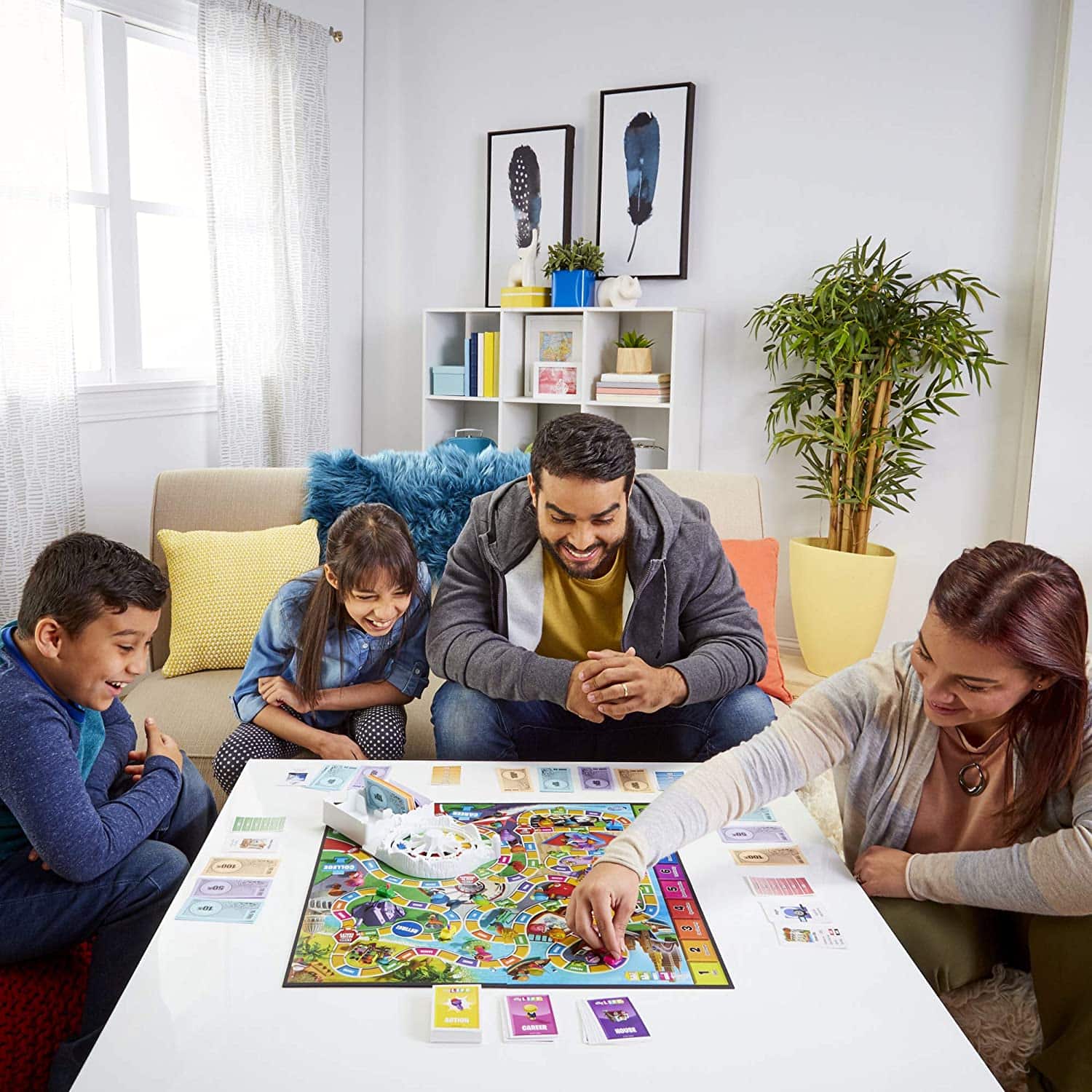 Game of Life 2021 'Your Life Your Way' Total Product Offering with game board, money, cards, and spinner, in a lifestyle photoshoot with family in their home. Made by Hasbro Gaming