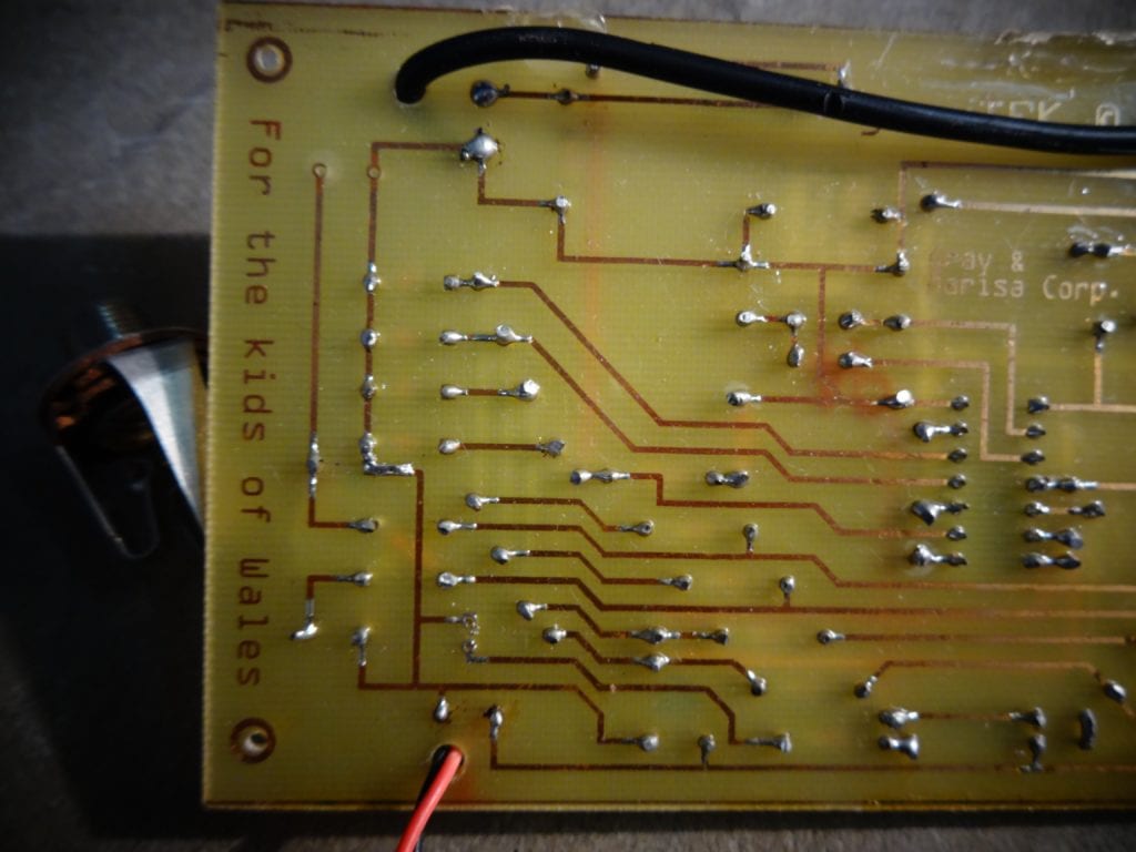 Gray Bright circuit board with smily face of components that is a PONG game in a circuit board with battery holder and buttons close up of components back of PCB on the left side